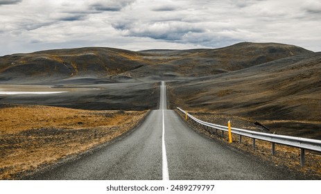 A long, empty road stretching through a barren, hilly landscape under a cloudy sky. Iceland - Powered by Shutterstock