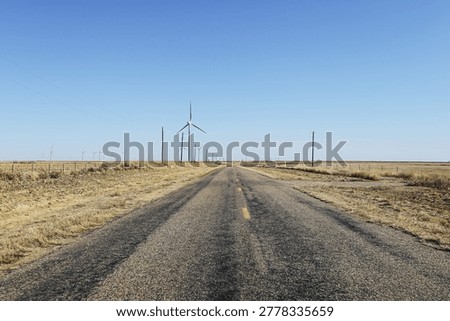 Long empty road through dry landscape with wind turbines on horizon. Renewable energy infrastructure in rural countryside. Transportation and sustainable power concept.