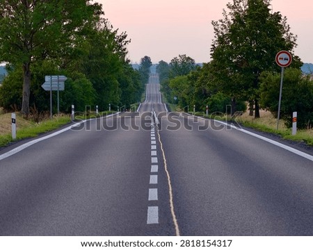 Long empty two-lane rural road curves over gentle hills at dusk, lined with dense green trees and roadside posts, white center and edge markings visible, soft pink glow across horizon