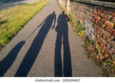 Long Evening Shadows Of A Man And Woman Walking The Dog On A Leash At Golden Hour Dusk. They Are Walking On A Pavement Next To A Grass Verge And An Interesting Textures Red Brick Wall.