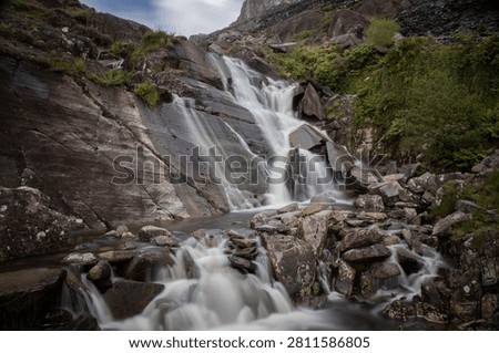 A long exposure captures the silky flow of a natural waterfall plunging over dark, ragged slate rocks and boulders in a rugged, green mountain landsca