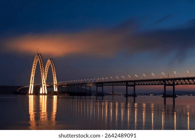 A long exposure captures a stunning cable-stayed bridge illuminated at twilight, its reflection shimmering on the calm water. The sky displays a beautiful gradient of colors.