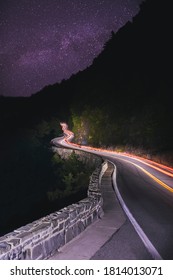 A Long Exposure Of Cars Passing Through A Winding Road On The Side Of A Mountain Under The Milky Way Sky At Night