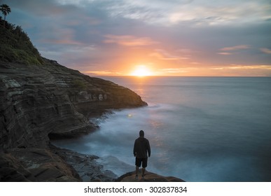 Long exposure creating a motion blur of sunrise from the cliffs on the east coast of Oahu, Hawaii overlooking the ocean. A man standing in the foreground looking down the coast toward the sun. 