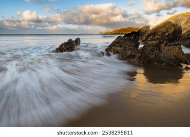 long exposure ocean scene on the dingle peninsula in ireland, showing movement of water at the shoreline, rocky coast, warm glow of sun on sand, and clouds.  - Powered by Shutterstock