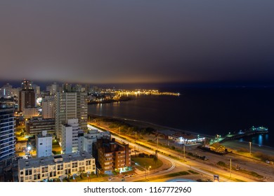 Long Exposure Panoramic Night View Of The Coastline At A Holiday Resort In Punta Del Este, Uruguay: The Quiet Empty Beach, The Calm Water, The Car Lights Passing And The Illuminated Harbor At The Back