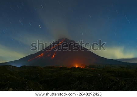 A long exposure photograph captures a Merapi volcano erupting at night, with lava flows cascading down its slopes. The night sky is filled with star trails, and the landscape glows.