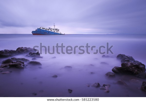 A long\
exposure of smooth serene ocean waters at a rocky beach and a\
capsized boat near shore after a hurricane at\
dusk