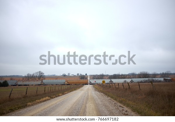 Long freight train crossing a remote rural dirt
road leading through farmland in a straight receding view under
grey cloud