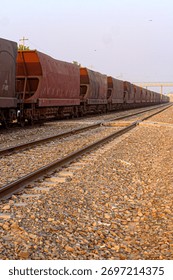 Long freight train of rust-colored hopper cars parked on parallel tracks, stretching into the distance over gravel under a hazy sky.

