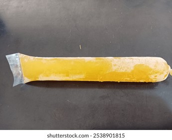 A long, frozen yellow ice pop in a clear plastic wrapper, lying flat on a dark surface. The ice pop has a smooth texture and a tapered end, typical of frozen treats.