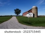 a long gravel driveway leads to a red barn, silo, and white outer building on a farm with a bright blue sky and a single tree in the background