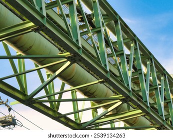 A long green industrial pipeline supported by metal framework stretches across a bright blue sky with light clouds, above buildings and power lines. - Powered by Shutterstock