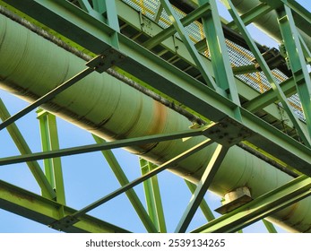 A long green industrial pipeline supported by metal framework stretches across a bright blue sky with light clouds, above buildings and power lines. - Powered by Shutterstock