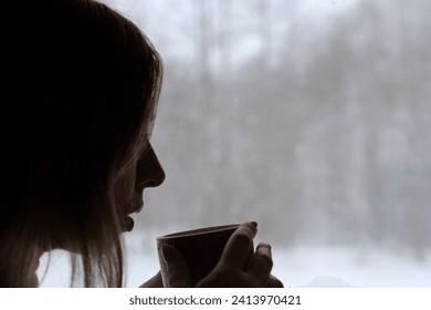 Long hair woman standing among the window and drinking hot tea from mug. Winter landscape. Lady drinks coffee looking at snowfall. Depression seasonal affective disorder female sad alone. Flu season