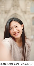 Long Haired Asian Girl Smiling While Taking Selfie Against The Backdrop Of Old Architecture. Pretty Japanese Woman Making Content For Her Blog In City