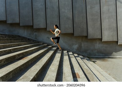 Long haired Asian woman in tracksuit runs up stone stairs past wall with concrete panels training on city street side view - Shutterstock ID 2060204396