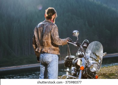 Long Haired Biker Standing Near His Custom Made Cruiser Motorcycle, Wearing Leather Jacket And Blue Jeans. Looking Into Distance. Shot From The Back.