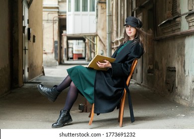Long haired girl in black coat, cap and green dress reading book on vintage chair in backstreet with shabby walls