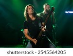 Long haired guitarist playing electric guitar solo at live rock concert with bandmate singing, illuminated by green stage lights for energetic performance