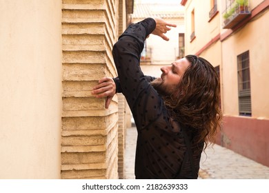 Long Haired Man Dancing Flamenco With Black Shirt And Red Roses. He Makes Dancing Postures With His Hands In A Typical Narrow Street Of Seville. Flamenco Dance Concept Cultural Heritage Of Humanity.