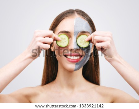 Long haired woman in clay mask holding slices of cucumber against her eyes