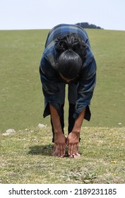 A Long Haired Young Man Doing Warm Up By Uttanasana Yoga Pose (Standing Toe Touch) Stretch Exercise In The Nature.