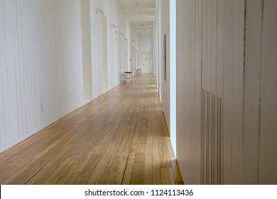 A Long Hallway With Birch Hardwood Floors And Pure White Walls. There's Windows Along One Wall And The Sun Is Shining Through Casting Shadows On The Floor. At The End Of The Hall Is A Single Bench. 