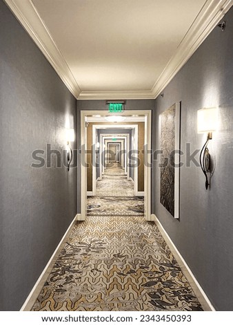 long hallway with gray walls and a carpeted floor. The hallway is well-lit by a series of chandeliers that hang from the ceiling. There are several doors along the hallway.