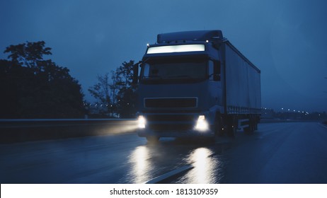 Long Haul Semi-Truck with Cargo Trailer Full of Goods Travels At Night on the Freeway Road, Driving Across Continent Through Rain, Fog, Snow. Industrial Warehouses Area. Front Shot