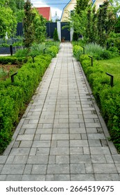 Long, inviting paved pathway bordered by manicured green hedges and lush garden beds, leading towards distant houses under bright sky, showcasing well-maintained backyard landscape