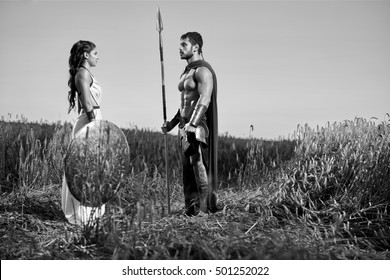 Long Journey Ahead. Monochrome Shot Of A Beautiful Greek Woman Standing In The Field With Her Man Handsome Strong Spartan Warrior Holding His Shield Copyspace 