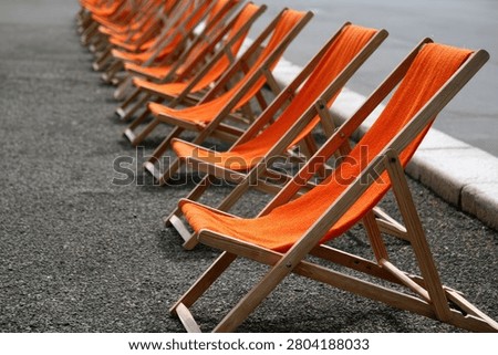 A long line of empty orange deck chairs are arranged on a gravel surface next to a body of water.