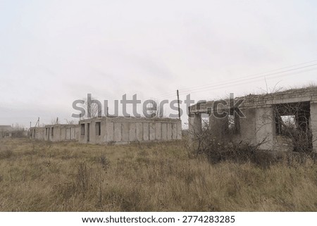 A long line of identical windowless roofless concrete building shells — never completed after the Chornobyl disaster — stretches across a flat rural Ukrainian landscape, overgrown with dry autumn grass under a heavy grey sky.