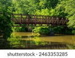 a long metal rust colored bridge over the silky brown waters of Little River surrounded by lush green trees, grass and plants with blue sky and clouds at Olde Rope Mill Park in Woodstock Georgia	