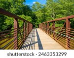 a long metal rust colored bridge over the silky brown waters of Little River surrounded by lush green trees, grass and plants with blue sky and clouds at Olde Rope Mill Park in Woodstock Georgia	