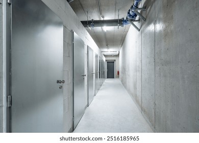 A long, narrow, concrete corridor with metallic doors, industrial lighting, and no visible people, leading towards a closed exit door at the far end. - Powered by Shutterstock