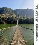 A long, narrow suspension bridge stretches across a calm lake, with lush green hills and a clear blue sky in the background. The image captures a sense of adventure and exploration.