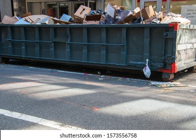 Long Overflowing Metal Dumpster On A Asphalt Street In Front Of Businesses