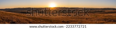 Long panorama of a sunset over a golden autumn prairie. The small hills are highlighted by the late day sun. The sun appears as a sunstar.
