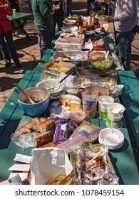 A Long Picnic Table Full Of Food At An Outdoor Picnic Potluck Community Lunch With A Few People Standing Around In The Background.