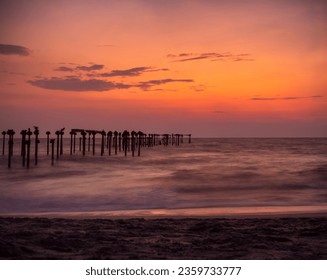 Long pier extending into the ocean at sunset. The pier is made of wood and has a long, curved shape. The waves are crashing against the shore and the sky is a vibrant orange and pink. - Powered by Shutterstock