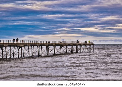 A long pier extends over a choppy sea under a cloudy sky.  Several people are visible on the pier, seemingly fishing. The overall color palette is muted blues and grays in Saltburn-by-the-Sea, UK. - Powered by Shutterstock