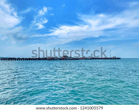 A long pier to the ocean between the blue sea and blue sky. There are many cars in line to get in the ferryboat. The great view from the shore of the island.