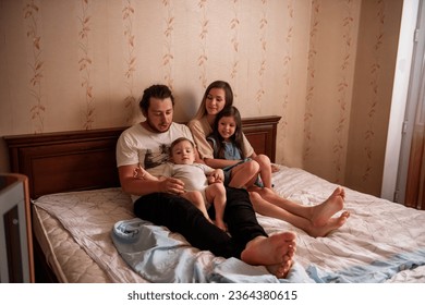 Long range shot of beautiful family on the bed in the morning in the sun from window. Tactile hugs of mother, father, daughter, son. Family weekend, relaxation at home. General view of family bedroom - Powered by Shutterstock