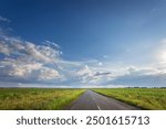 A long road with a clear blue sky above it. The sky is dotted with clouds, but the sun is shining brightly. The road is empty, with no cars or people in sight. The scene is peaceful and serene