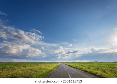 A long road with a clear blue sky above it. The sky is dotted with clouds, but the sun is shining brightly. The road is empty, with no cars or people in sight. The scene is peaceful and serene - Powered by Shutterstock