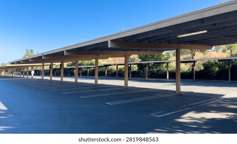A long row of empty covered parking spaces under a large carport structure with support pillars next to green trees on a bright sunny day under a clear blue sky