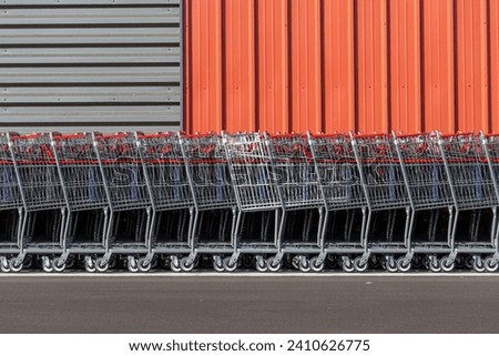 A long row of new metal shopping carts with red handles. The retail pushcarts are in front of an orange and grey colored metal outside wall of a shopping mall. The trolly wagons have small wheels.