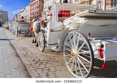 A long row of white carriages on a cobbled street of an old European city awaits tourists for a horse ride on a sunny day in Krakow, Poland